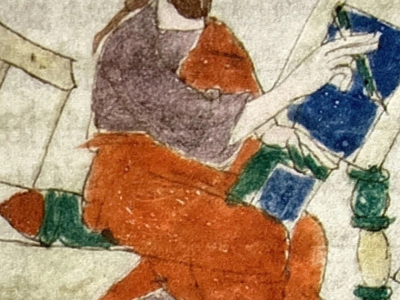 Uomo barbuto con capelli lunghi in veste rosso-viola seduto su una panca, tiene un libro aperto e punta il dito sulle pagine.