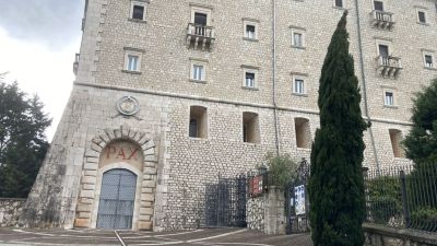 Ingresso laterale dell’abbazia di Montecassino, con il portale ad arco sormontato dalla scritta PAX in rosso; la facciata è in pietra chiara con piccole finestre e balconcini, sulla destra si vede un alto cipresso e l’accesso recintato.
