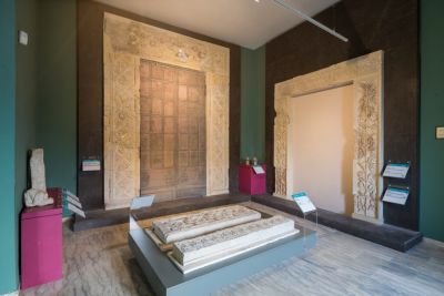 Interno del Museo dell’Abbazia di Montecassino con due grandi portali decorati esposti a parete, uno con battenti bronzei e cornice scolpita e l’altro con cornice ornamentale chiara; in primo piano lastre marmoree intagliate esposte su una piattaforma.