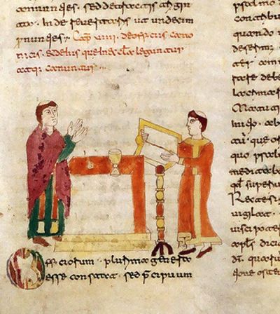 Miniatura medievale con due figure maschili: a sinistra un uomo in tunica verde e mantello viola con le mani alzate in gesto di discorso; a destra uno scriba in tunica arancione che regge un grande libro aperto su un leggio dorato. Tra i due, un altare quadrangolare coperto da un panno arancione con sopra un calice. In basso, un’iniziale miniata ornata.