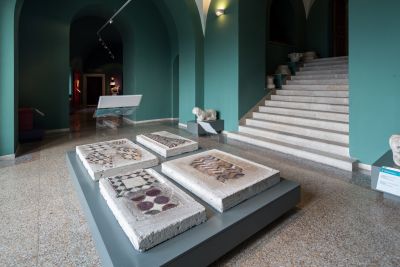Interno del Museo dell’Abbazia di Montecassino con pavimenti cosmateschi originali esposti su supporti in cemento, collocati al centro della sala verde acqua; sullo sfondo si vedono teche espositive, archi e una scala in marmo che sale al piano superiore.
