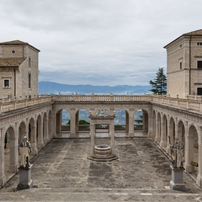 Cortile in pietra con pozzo centrale, archi e statue tra due edifici storici; sullo sfondo, montagne e cielo nuvoloso.