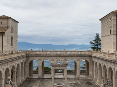 Cortile lastricato con un pozzo centrale, portici ad archi e statue, tra due edifici; sullo sfondo montagne e cielo nuvoloso.