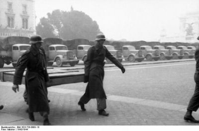 Soldati che trasportano un quadro imballato lungo una strada, con una fila di camion militari parcheggiati sullo sfondo.